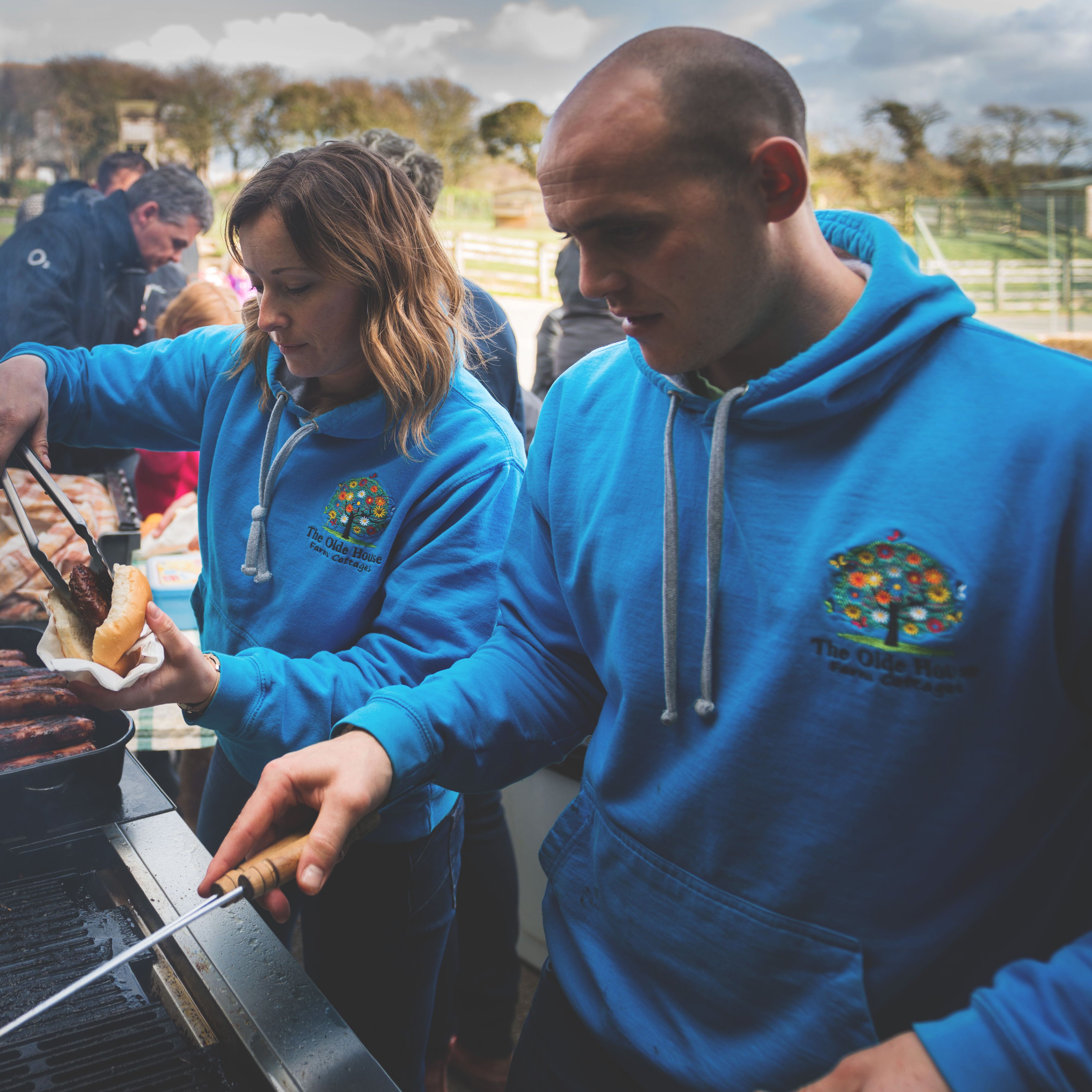 People wearing blue hoodies are grilling and serving food at an outdoor barbecue event, with a group of people and children waiting in the background.