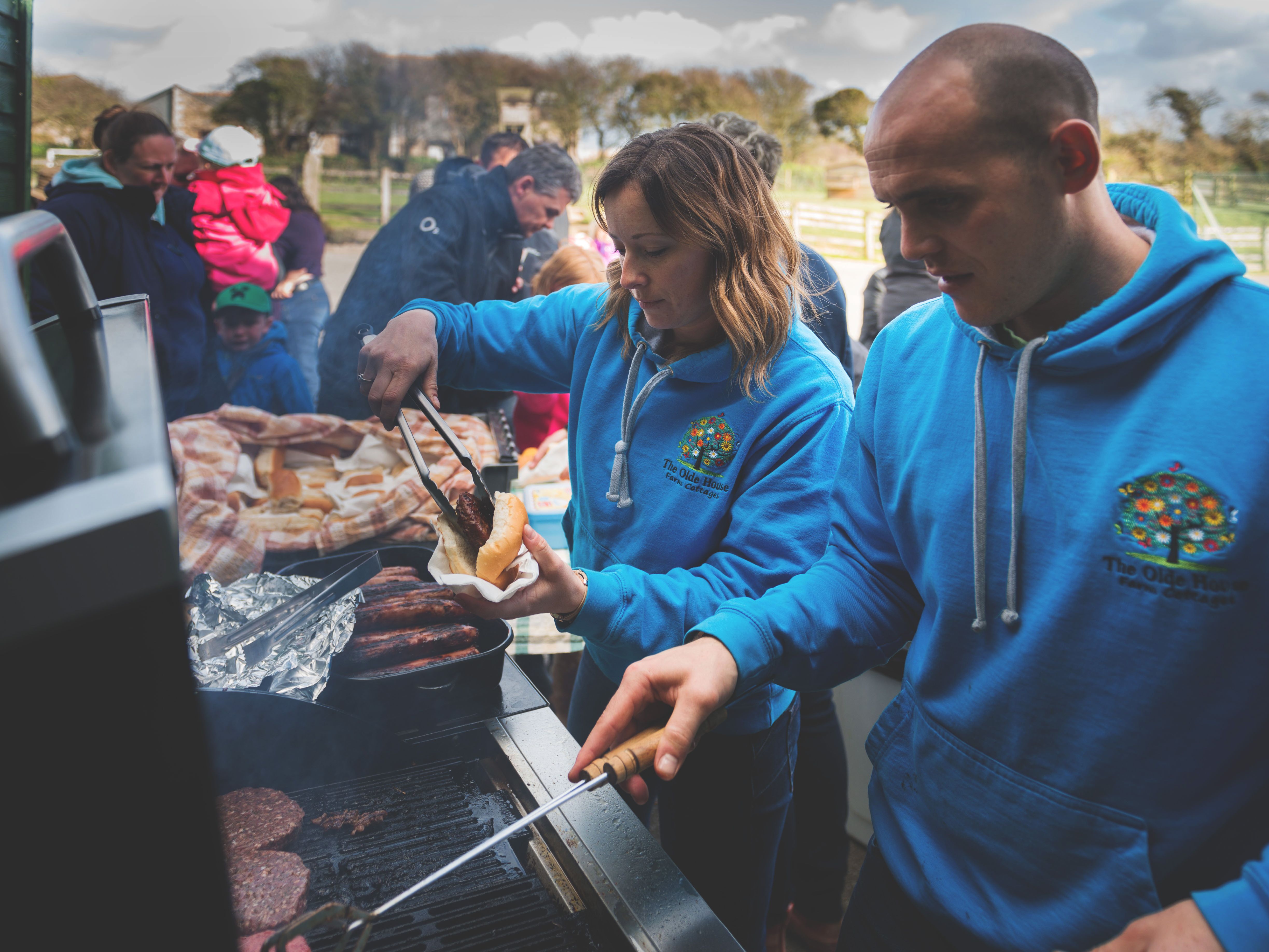 People wearing blue hoodies are grilling and serving food at an outdoor barbecue event, with a group of people and children waiting in the background.