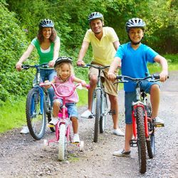Family with two adults and two children riding bicycles together on a forest path, all wearing helmets and smiling.