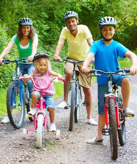 Family with two adults and two children riding bicycles together on a forest path, all wearing helmets and smiling.