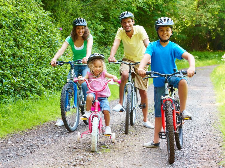 Family with two adults and two children riding bicycles together on a forest path, all wearing helmets and smiling.