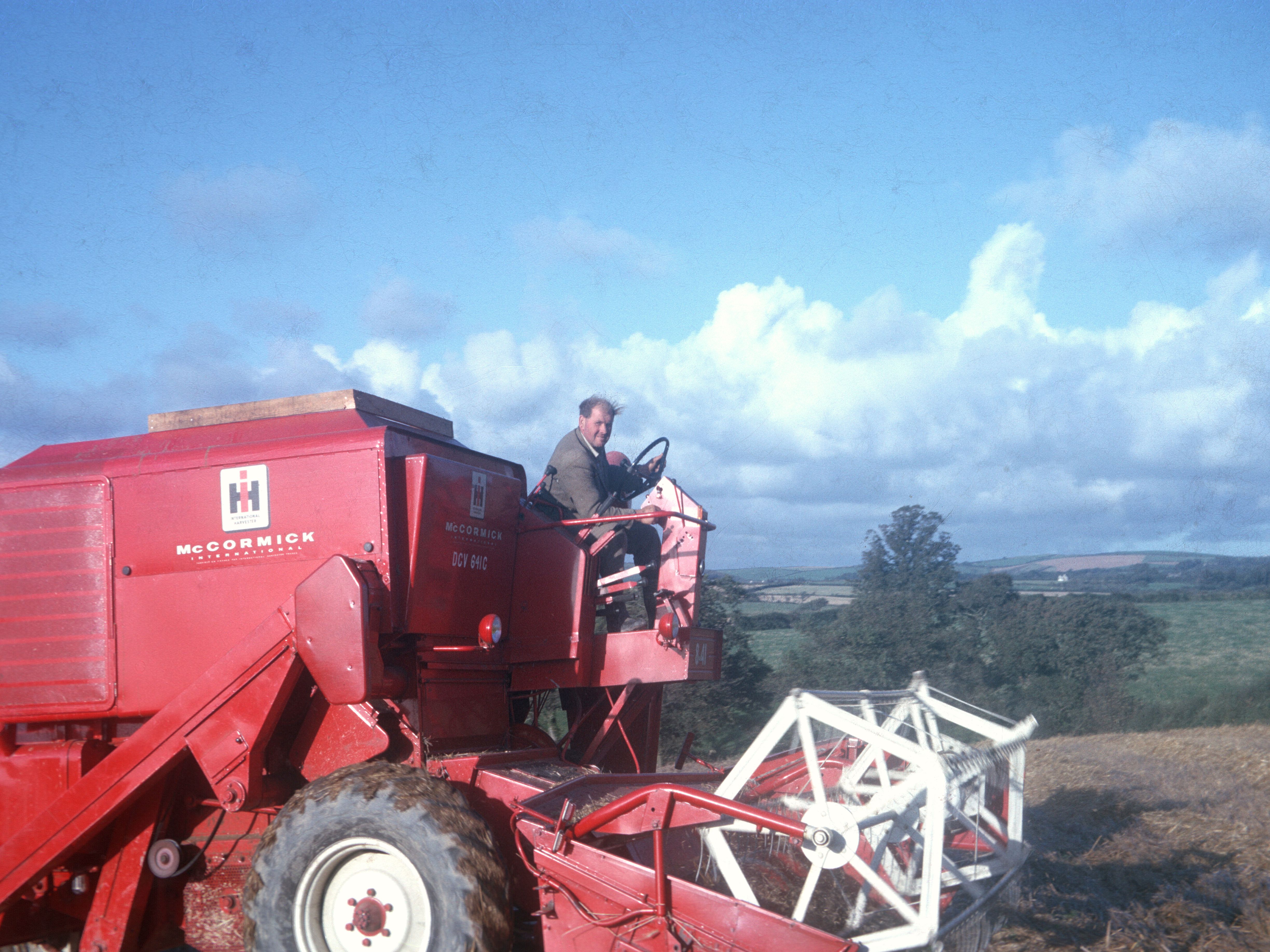 A man driving a red McCormick International combine harvester in a field with trees and hills in the background under a blue sky with clouds.