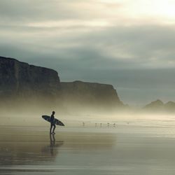A lone surfer walks along a misty beach near rocky cliffs, holding a surfboard under a cloudy sky.