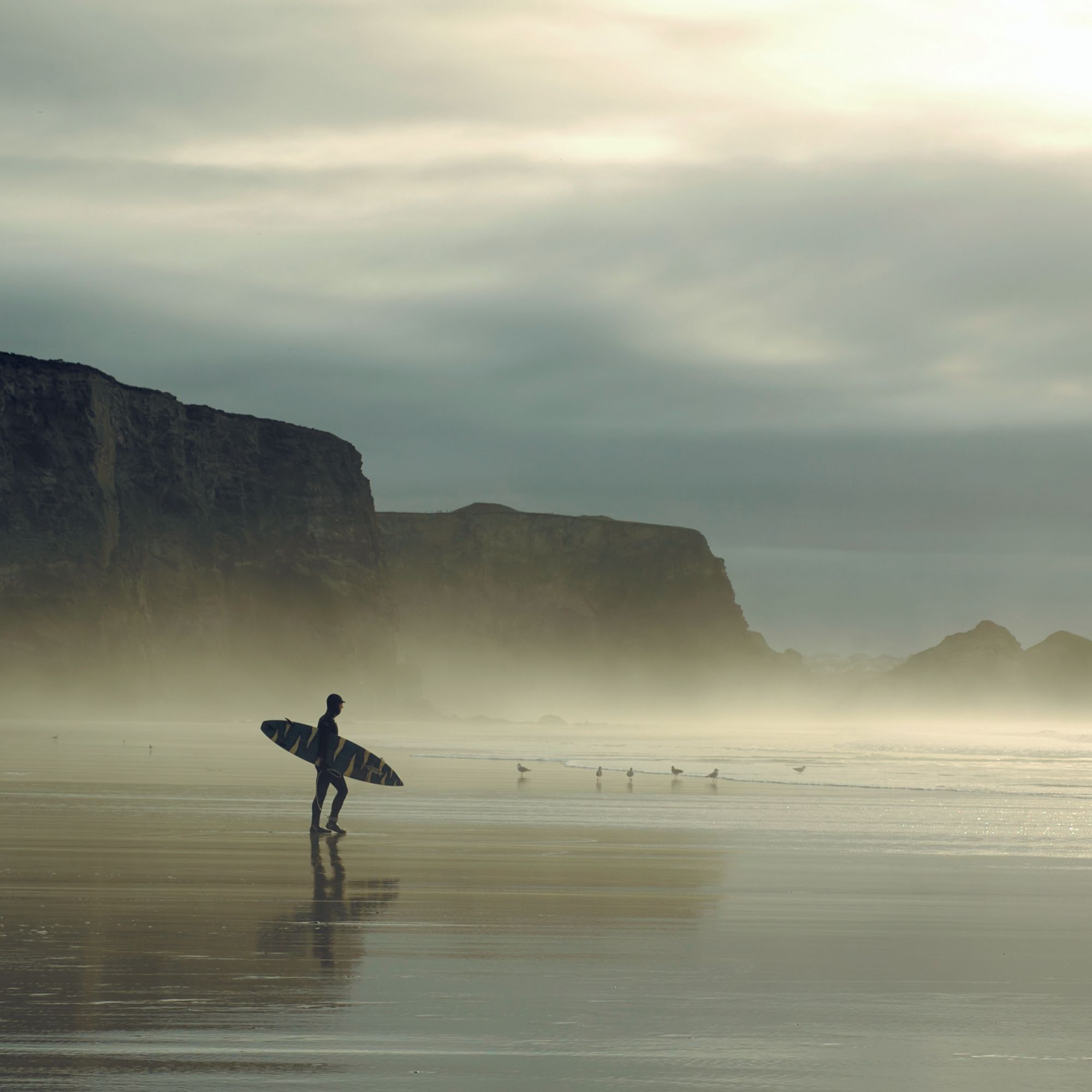 A lone surfer walks along a misty beach near rocky cliffs, holding a surfboard under a cloudy sky.