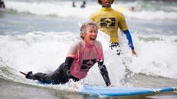 Older woman learning to surf, smiling and lying on a surfboard in the ocean, with an instructor standing nearby.
