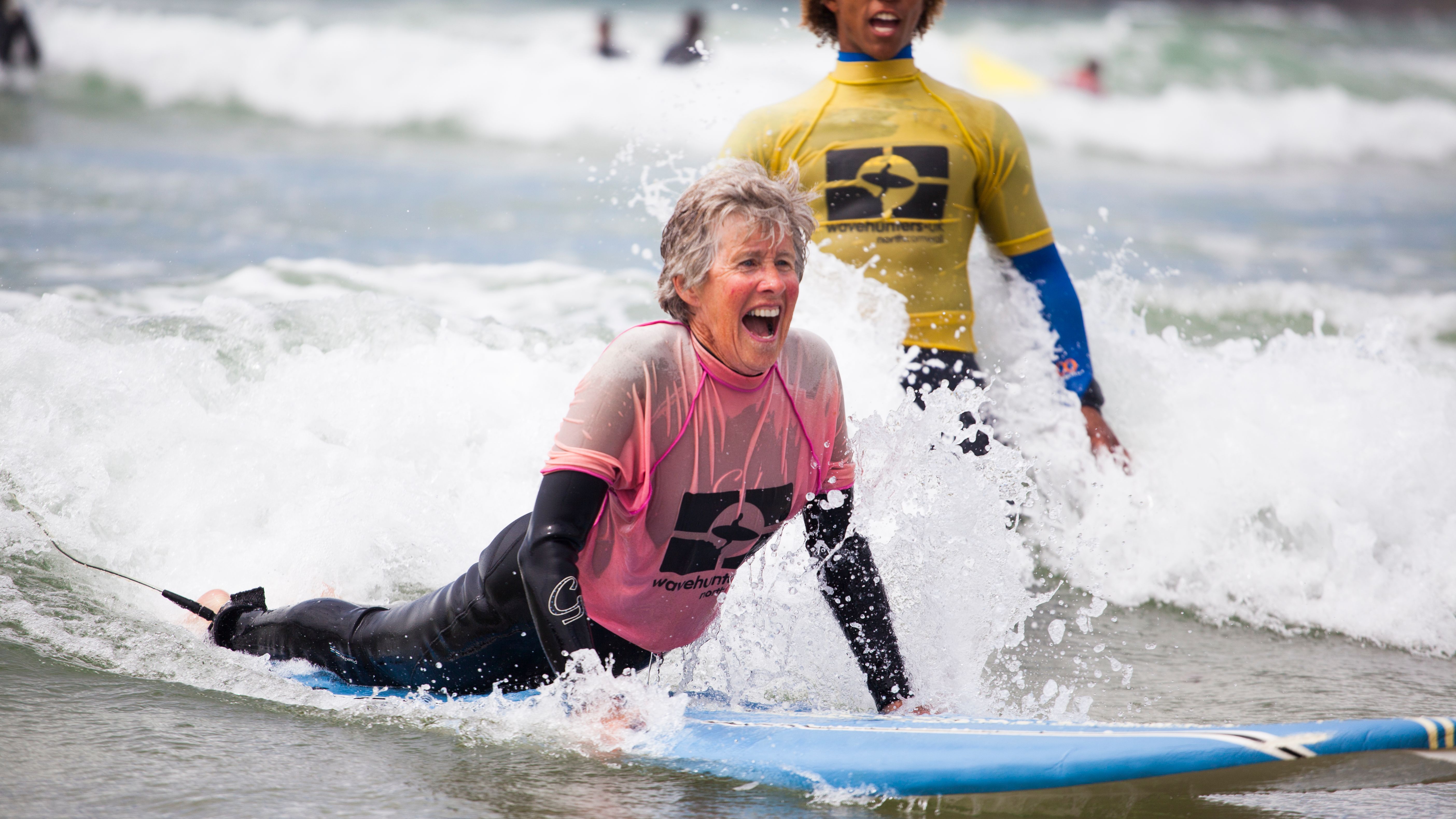 Older woman learning to surf, smiling and lying on a surfboard in the ocean, with an instructor standing nearby.