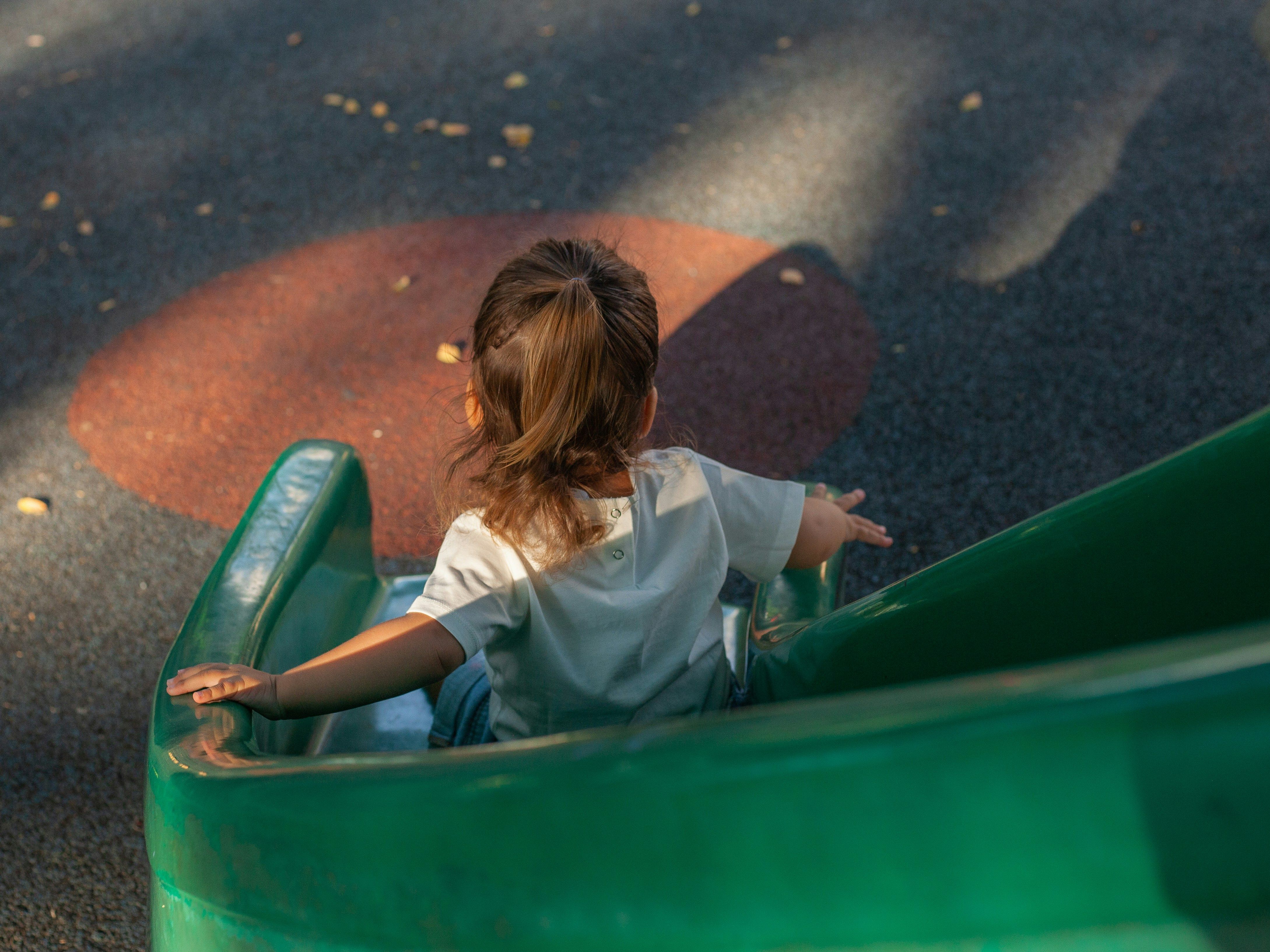 Child with brown hair in a ponytail wearing a white shirt, sliding down a green playground slide.