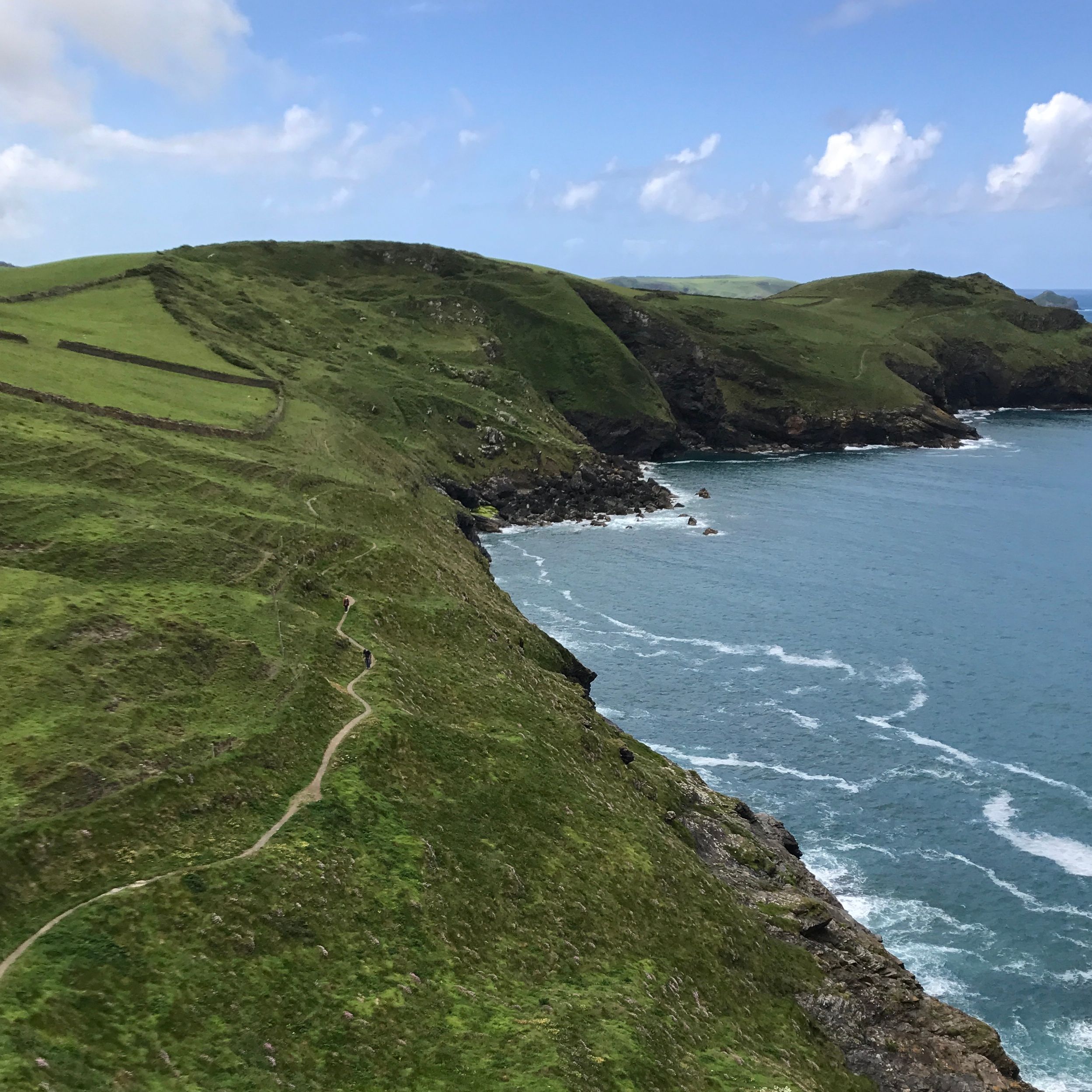 Coastal landscape with green hillsides, a winding path, and cliffs overlooking the blue ocean under a partly cloudy sky.