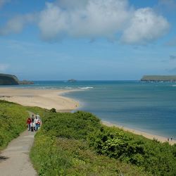 A scenic coastal landscape with a group of people walking on a path beside lush green bushes, overlooking a sandy beach and blue sea under a partly cloudy sky.