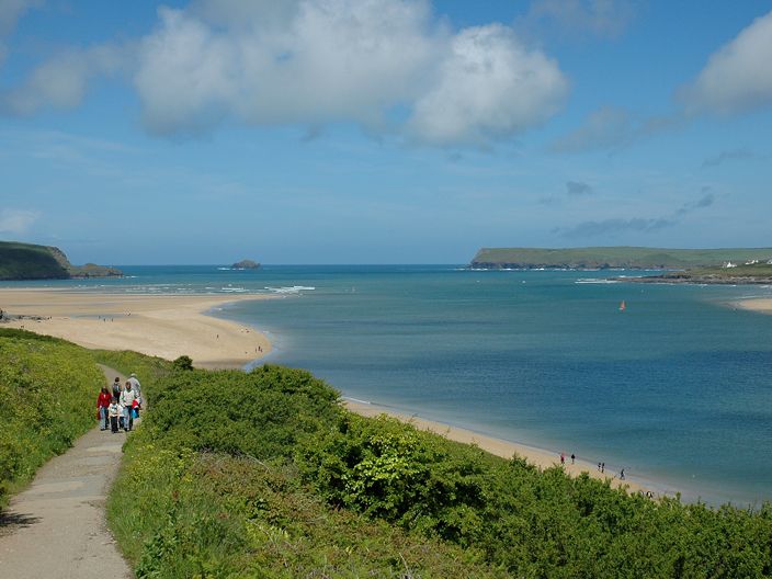 A scenic coastal landscape with a group of people walking on a path beside lush green bushes, overlooking a sandy beach and blue sea under a partly cloudy sky.