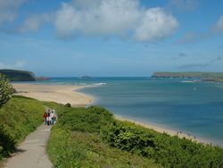 A scenic coastal landscape with a group of people walking on a path beside lush green bushes, overlooking a sandy beach and blue sea under a partly cloudy sky.