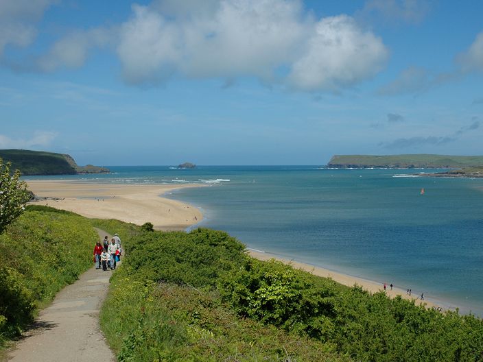 A scenic coastal landscape with a group of people walking on a path beside lush green bushes, overlooking a sandy beach and blue sea under a partly cloudy sky.