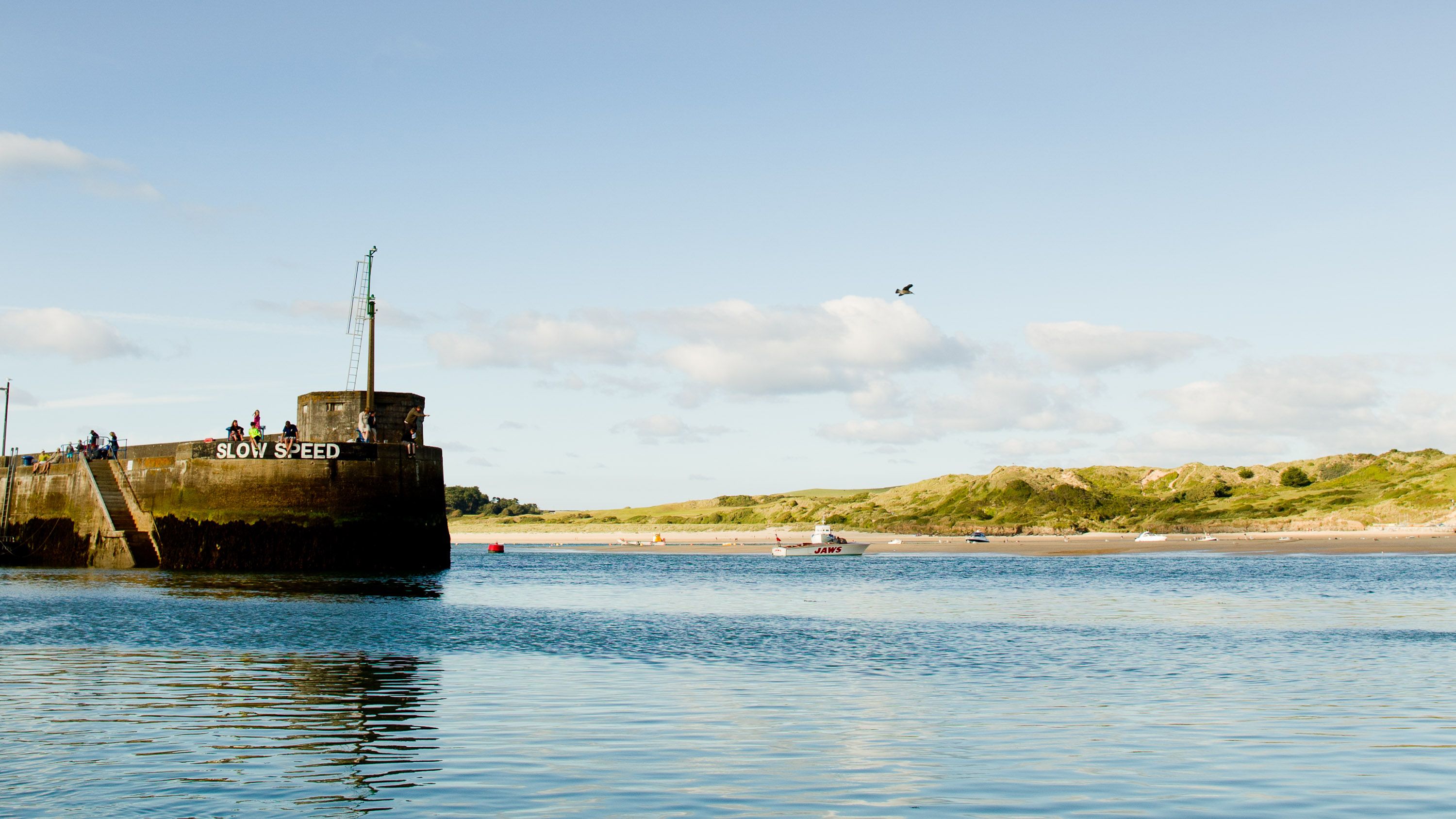 A calm harbor scene with a concrete pier labeled 'SLOW SPEED', clear blue water, a few people on the pier, grassy hills in the background, boats in the distance, and a partly cloudy sky.