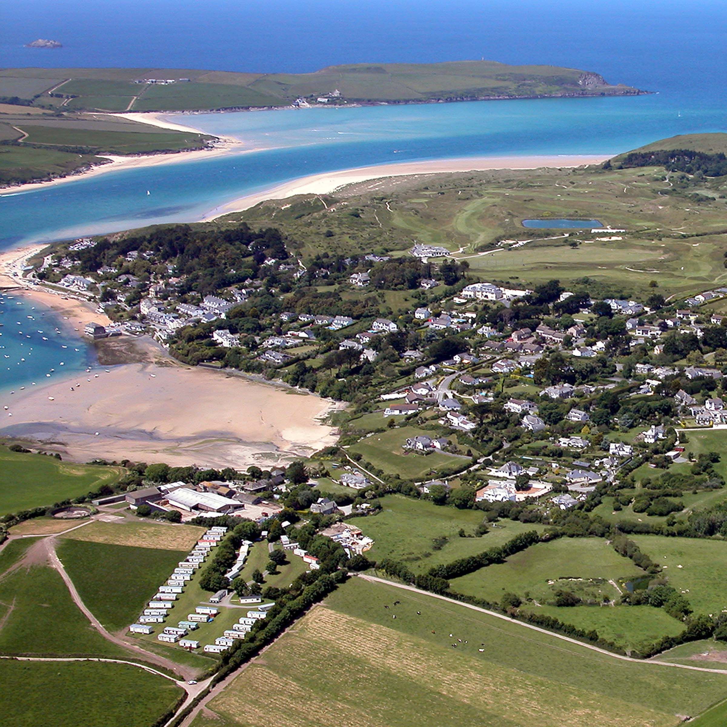 Aerial view of a coastal village with sandy beach, green fields, scattered houses, and a river flowing into the sea.