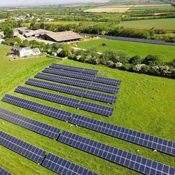 Aerial view of solar panels installed on a green field in a rural area, with a farm and buildings in the background surrounded by farmland and countryside.