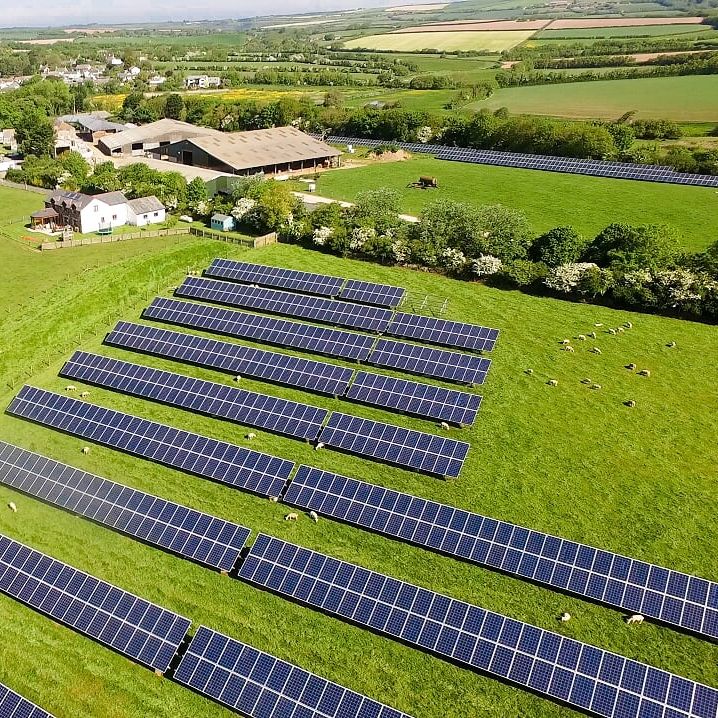 Aerial view of solar panels installed on a green field in a rural area, with a farm and buildings in the background surrounded by farmland and countryside.