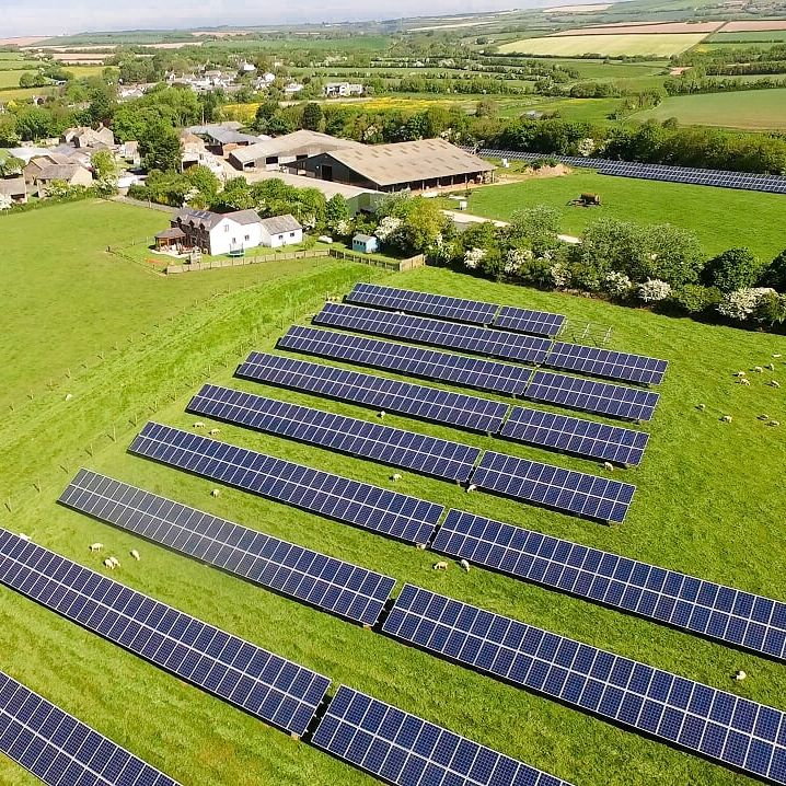Aerial view of solar panels installed on a green field in a rural area, with a farm and buildings in the background surrounded by farmland and countryside.
