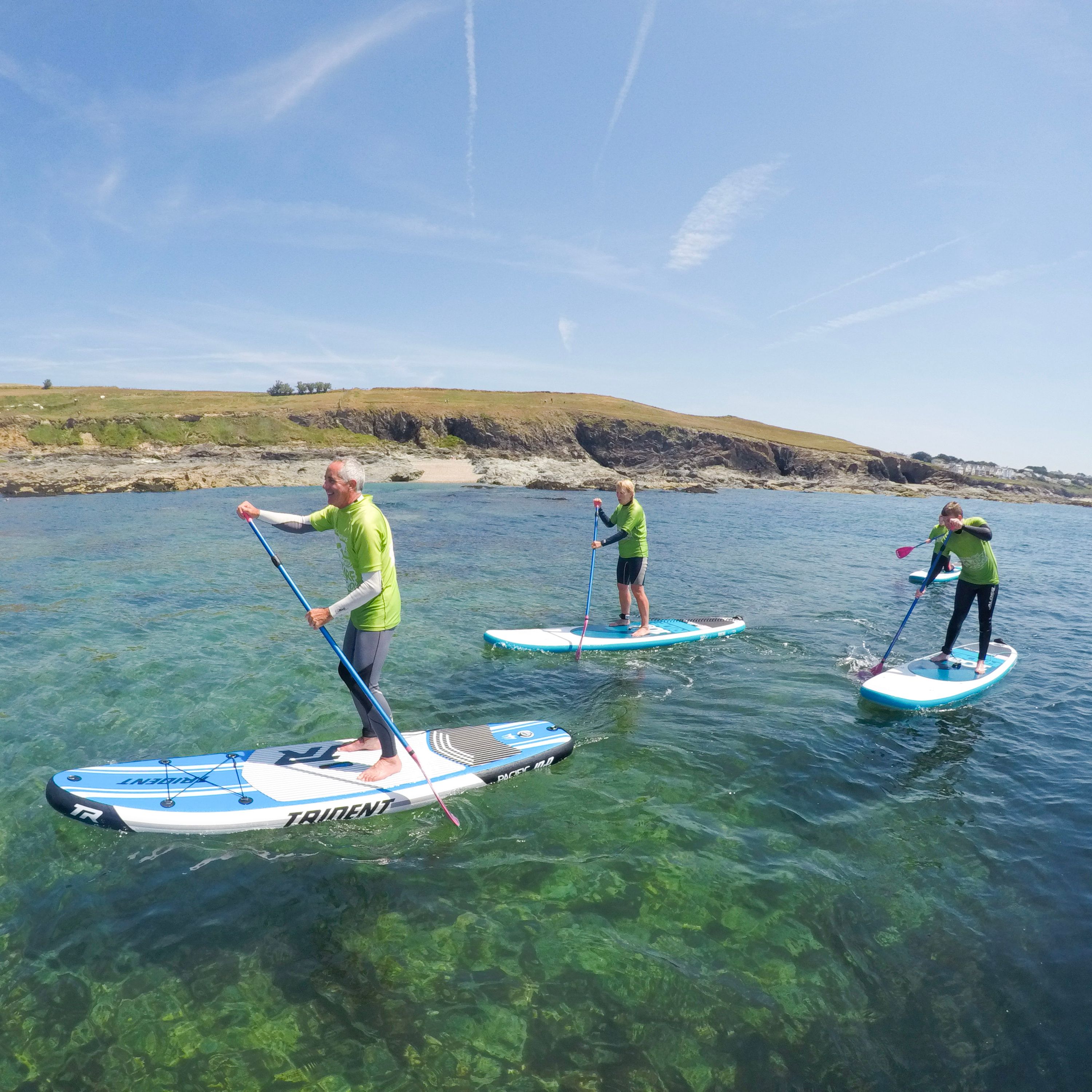 Three people paddle boarding on clear blue water near a rocky coastline under a sunny sky.