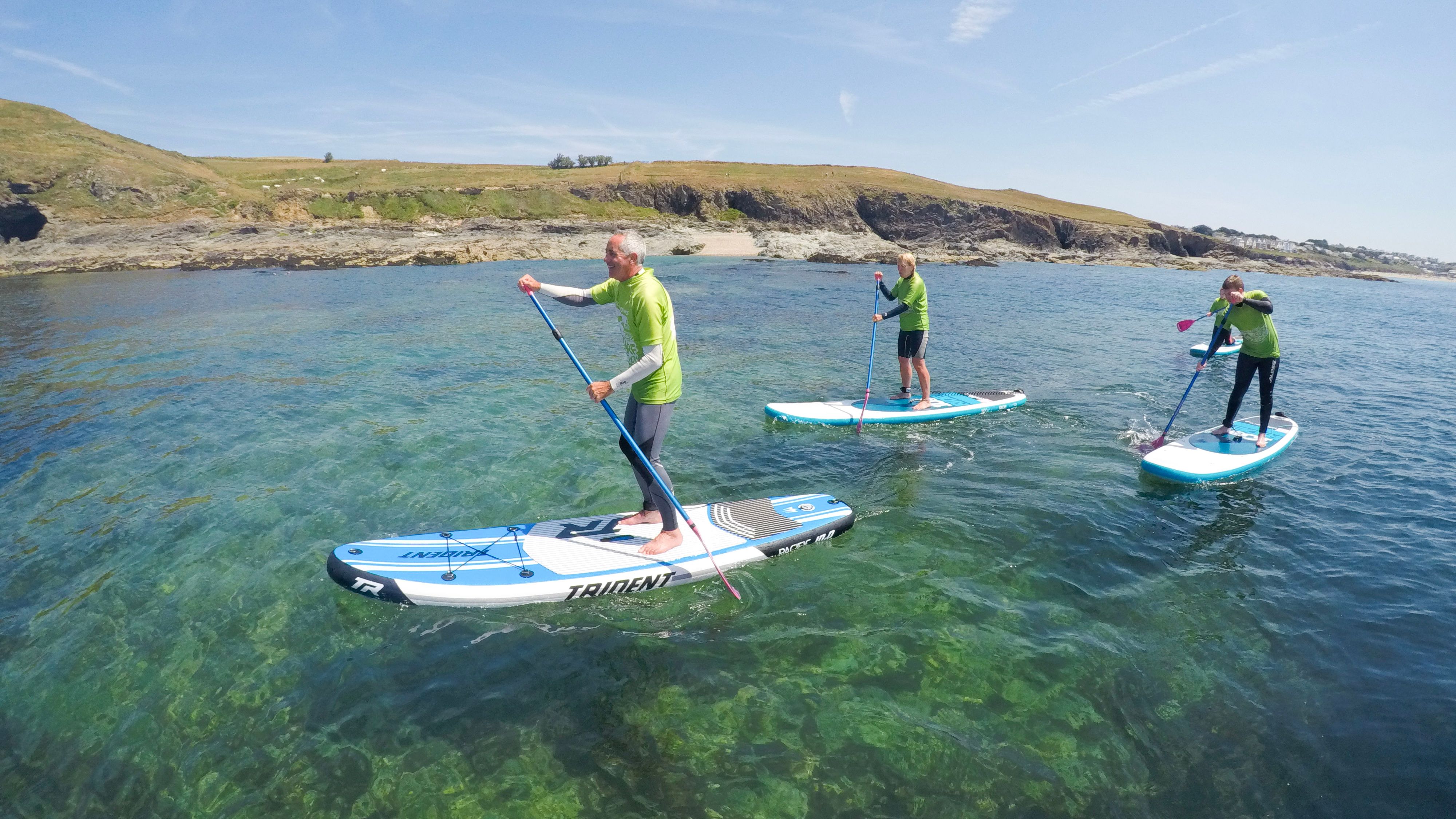 Three people paddle boarding on clear blue water near a rocky coastline under a sunny sky.