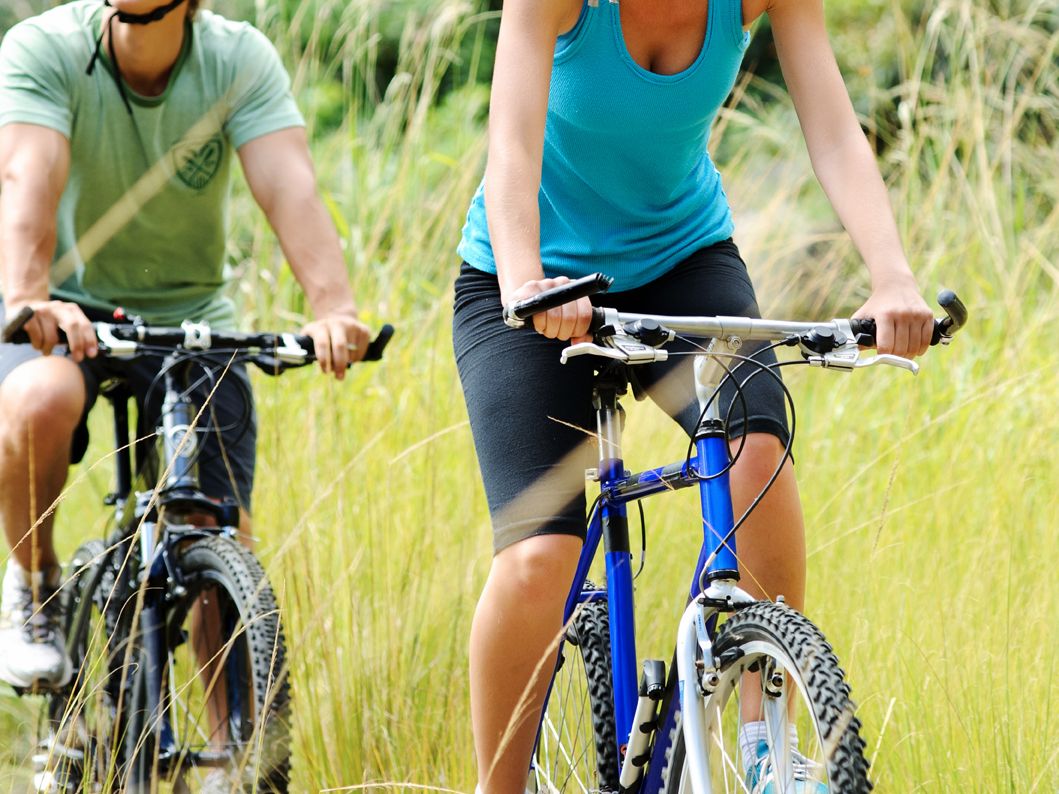 Two people riding mountain bikes on a grassy trail, wearing helmets and athletic clothing, surrounded by greenery.