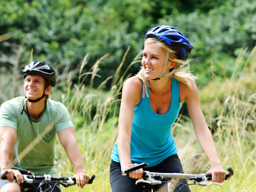 Two people riding mountain bikes on a grassy trail, wearing helmets and athletic clothing, surrounded by greenery.