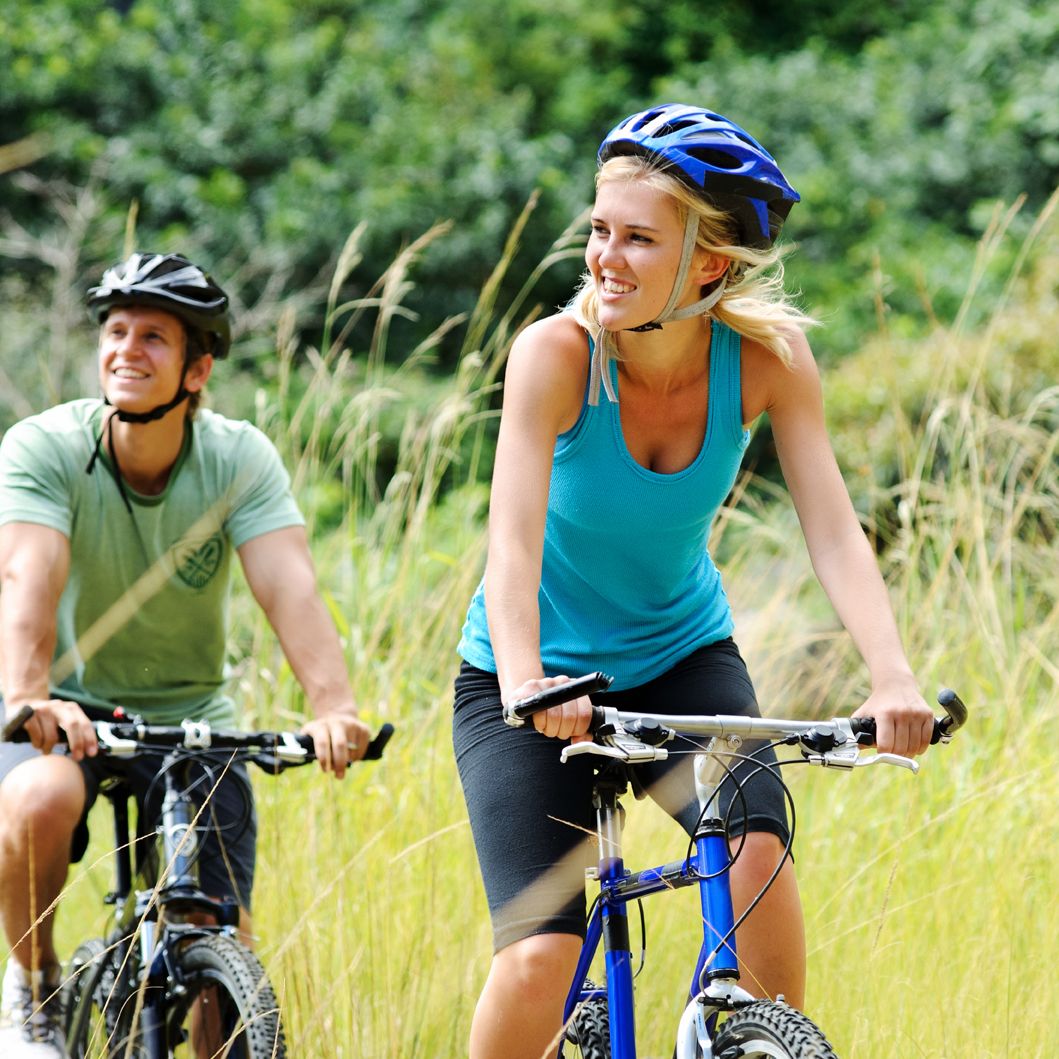 Two people riding mountain bikes on a grassy trail, wearing helmets and athletic clothing, surrounded by greenery.