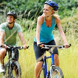 Two people riding mountain bikes on a grassy trail, wearing helmets and athletic clothing, surrounded by greenery.