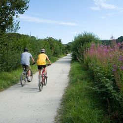 Two people riding bicycles on a gravel path surrounded by greenery and wildflowers under a blue sky.