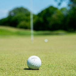 A close-up of a golf ball on a putting green with a flagstick and another golf ball in the blurred background on a sunny day.