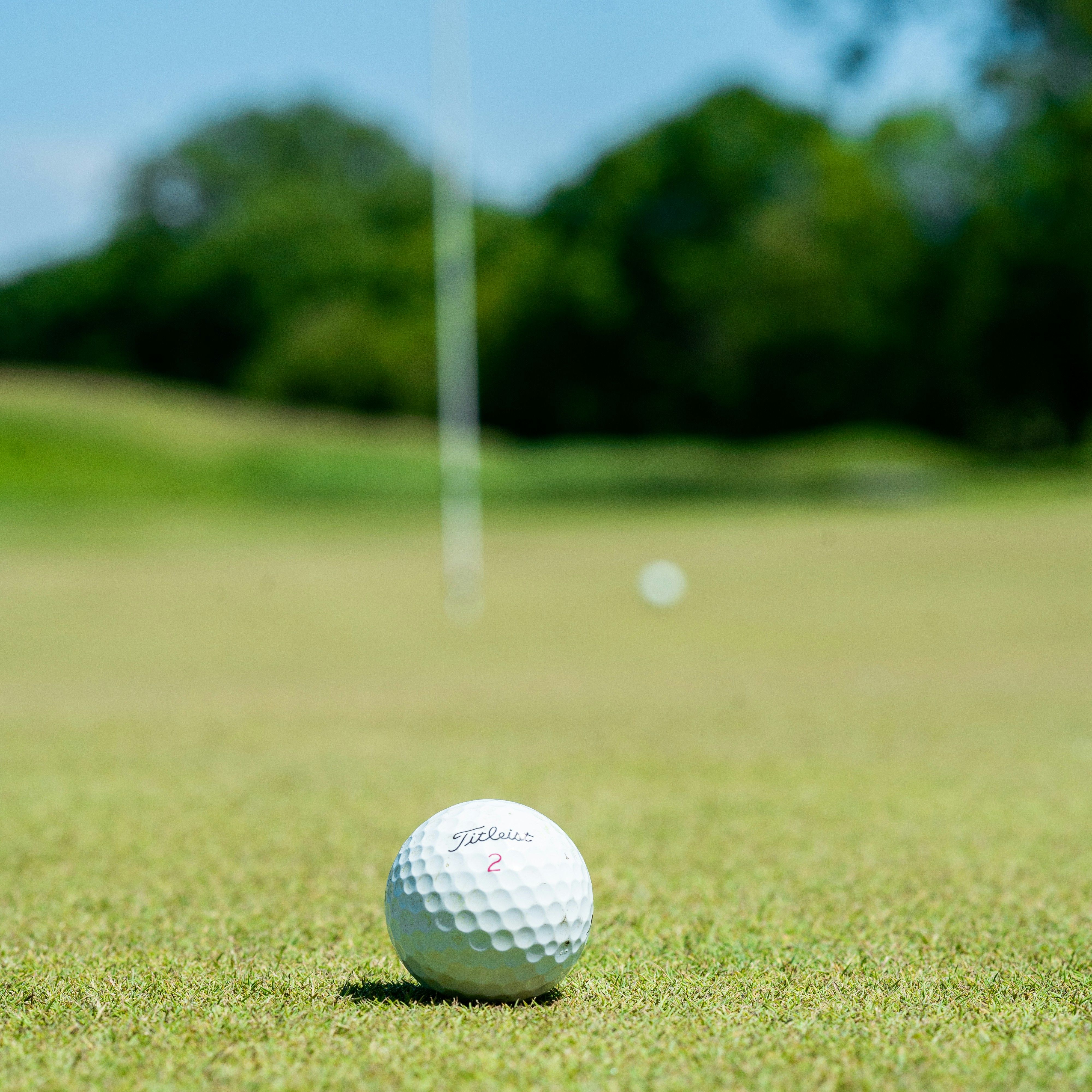A close-up of a golf ball on a putting green with a flagstick and another golf ball in the blurred background on a sunny day.