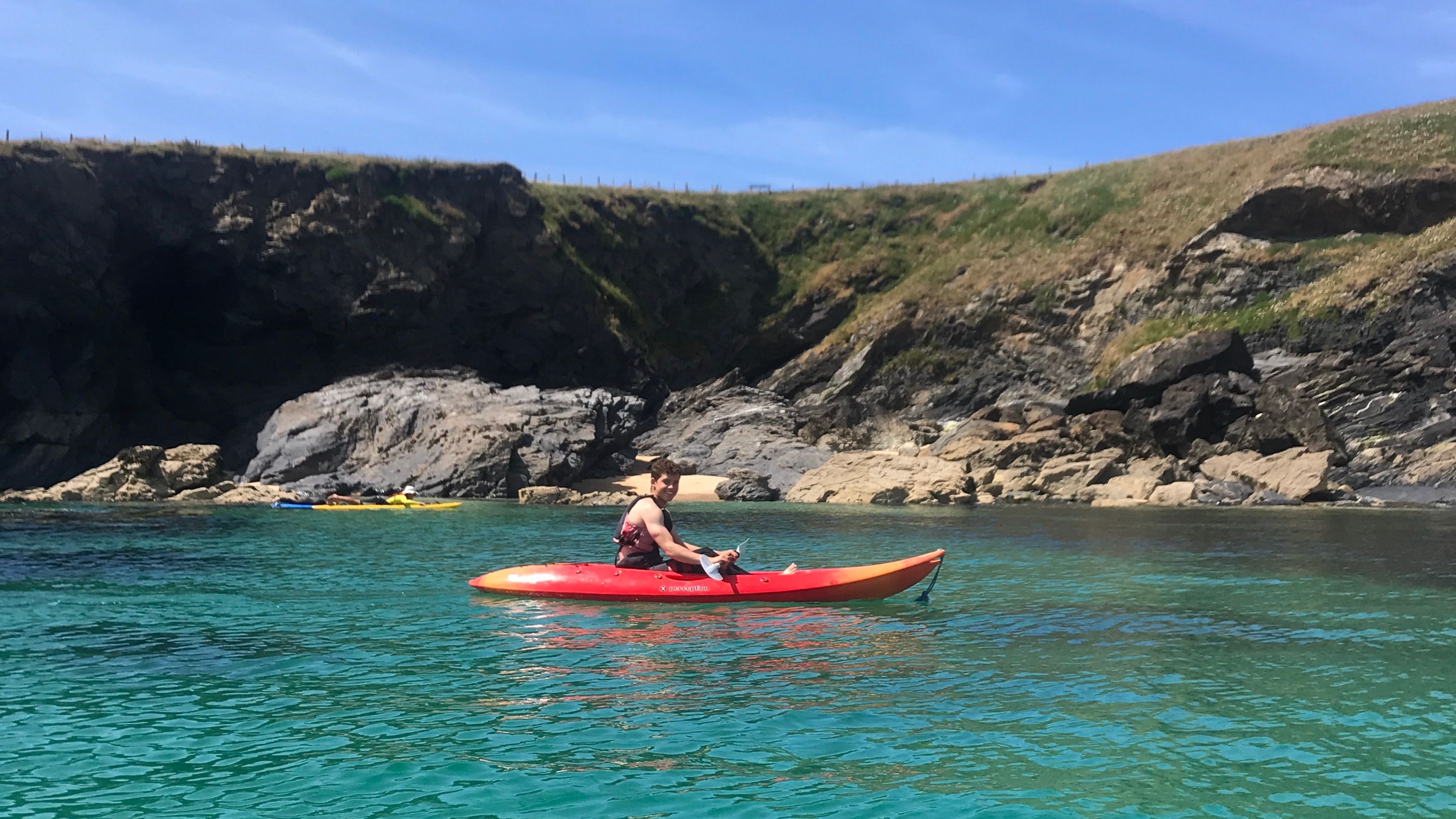 Person kayaking on clear turquoise water near rocky cliffs with grassy hills under a blue sky.