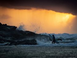 Surfer walking in the ocean near rocks with waves splashing at sunset with dramatic, cloudy sky.