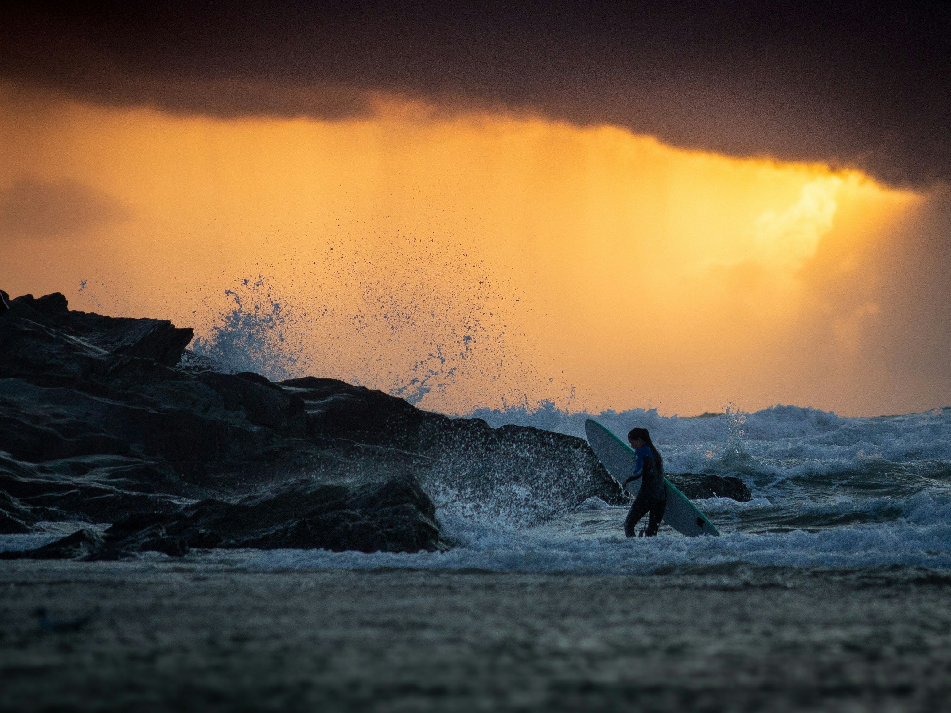 Surfer walking in the ocean near rocks with waves splashing at sunset with dramatic, cloudy sky.