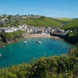 A picturesque seaside village with white houses perched on green hills, a sandy beach, and boats floating in a clear blue harbor under a bright sky.