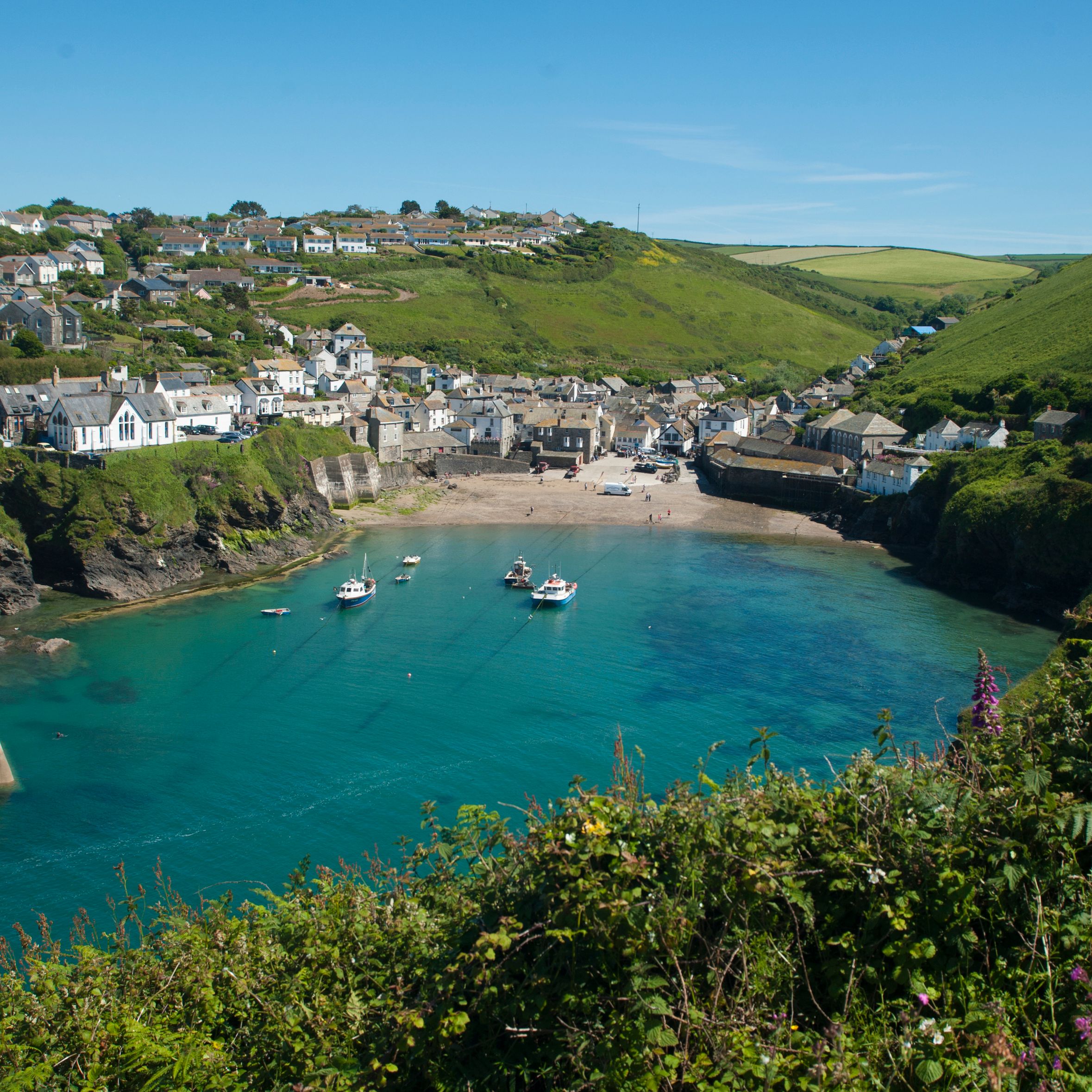 A picturesque seaside village with white houses perched on green hills, a sandy beach, and boats floating in a clear blue harbor under a bright sky.