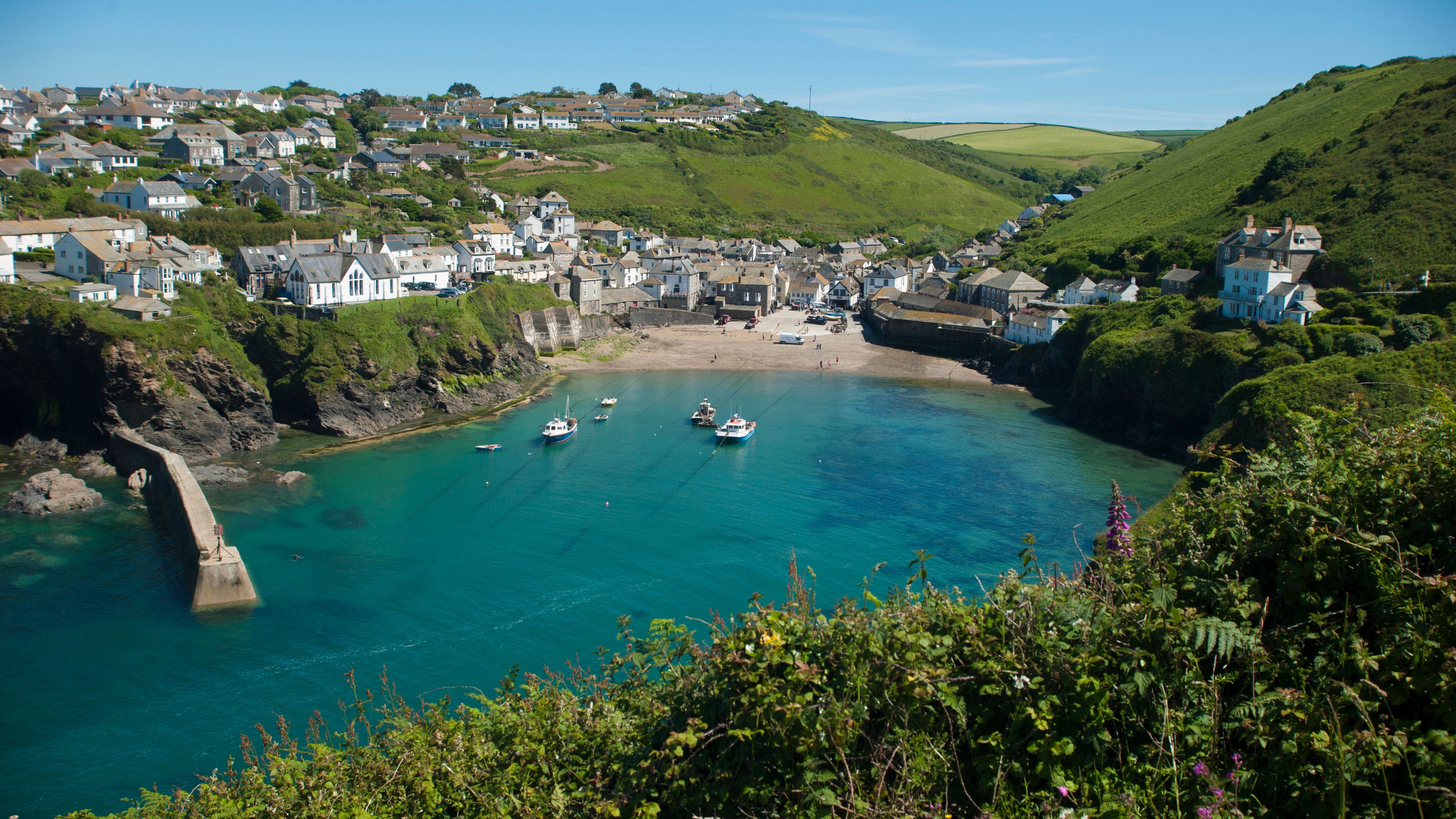 A picturesque seaside village with white houses perched on green hills, a sandy beach, and boats floating in a clear blue harbor under a bright sky.