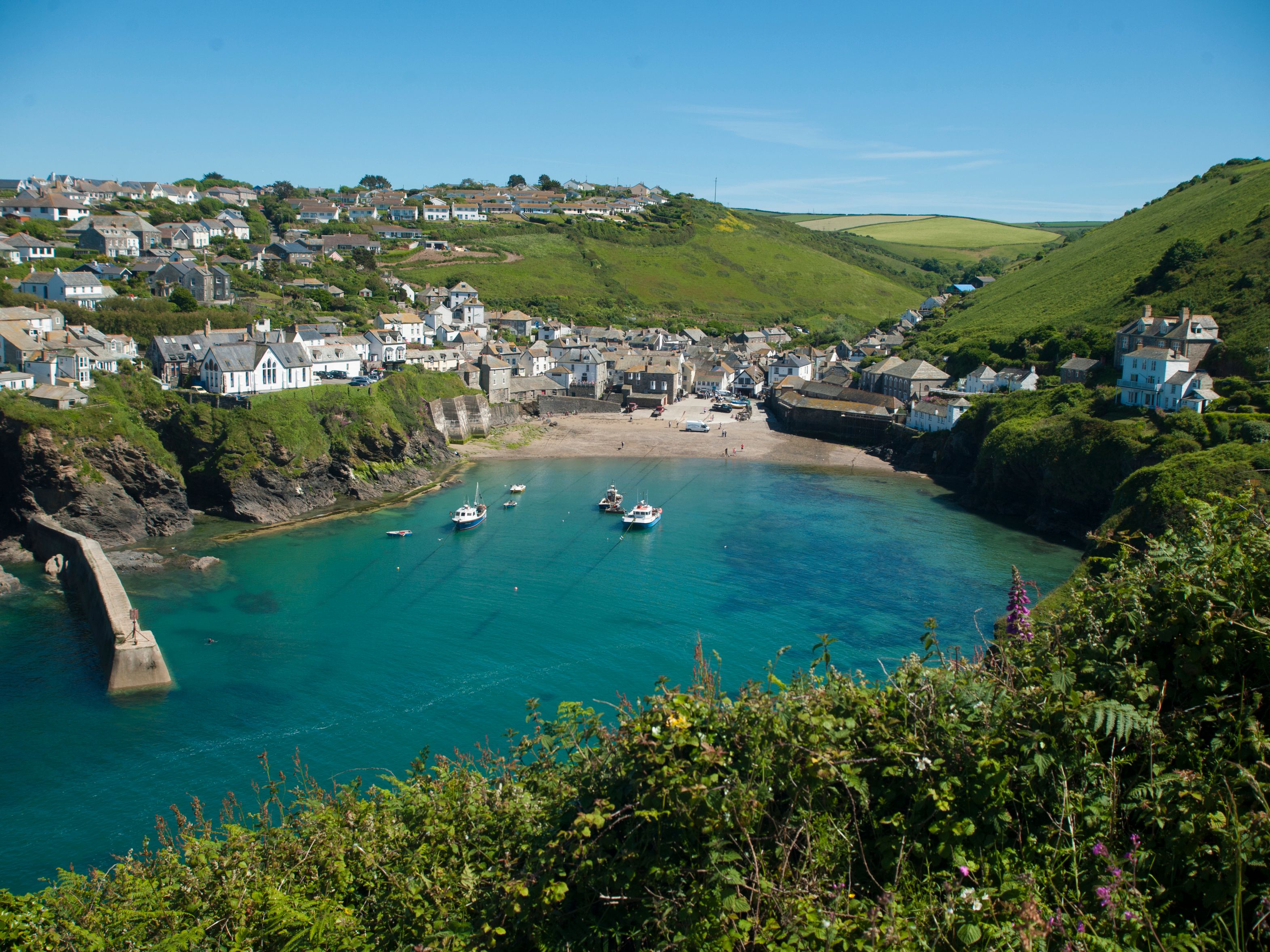A picturesque seaside village with white houses perched on green hills, a sandy beach, and boats floating in a clear blue harbor under a bright sky.