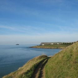 A coastal path running along grassy cliffs overlooking a calm blue sea, with distant houses and a rocky island visible under a clear sky.