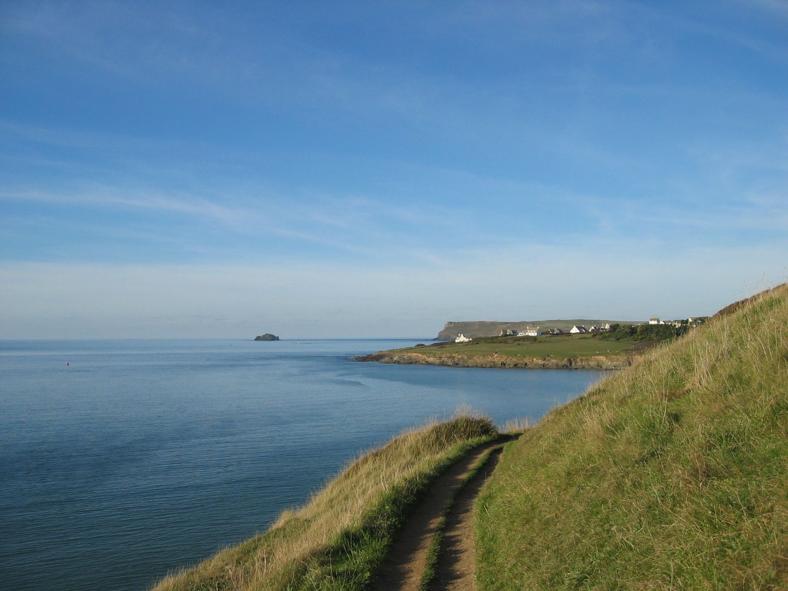 A coastal path running along grassy cliffs overlooking a calm blue sea, with distant houses and a rocky island visible under a clear sky.