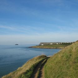 A coastal path running along grassy cliffs overlooking a calm blue sea, with distant houses and a rocky island visible under a clear sky.