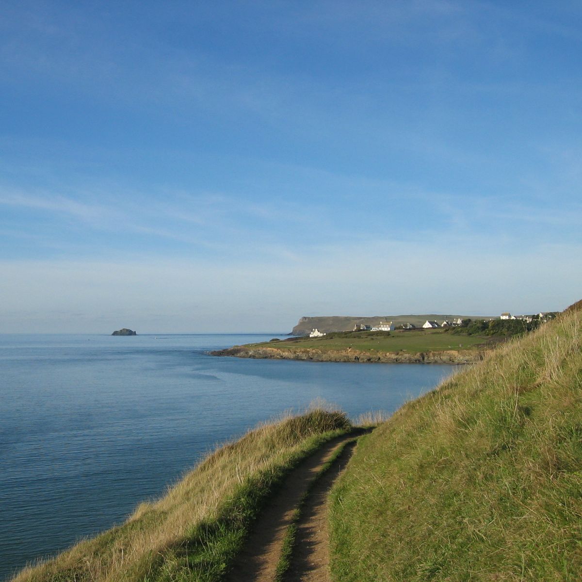 A coastal path running along grassy cliffs overlooking a calm blue sea, with distant houses and a rocky island visible under a clear sky.