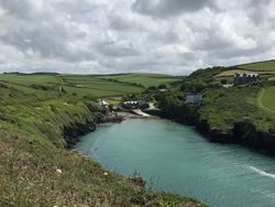 Scenic coastal inlet with turquoise water, rocky cliffs, green fields, and a small village with stone houses under a cloudy sky.
