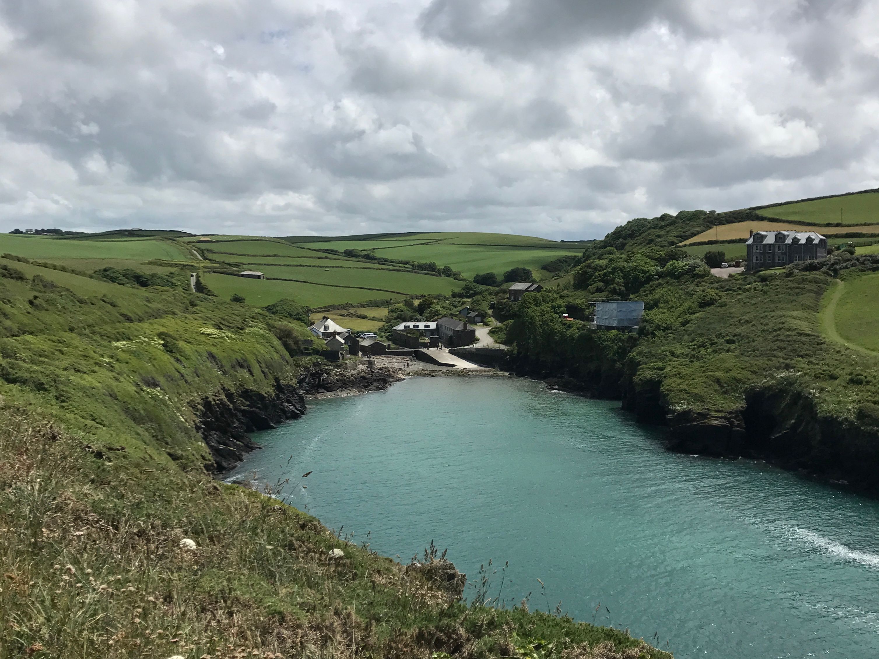 Scenic coastal inlet with turquoise water, rocky cliffs, green fields, and a small village with stone houses under a cloudy sky.
