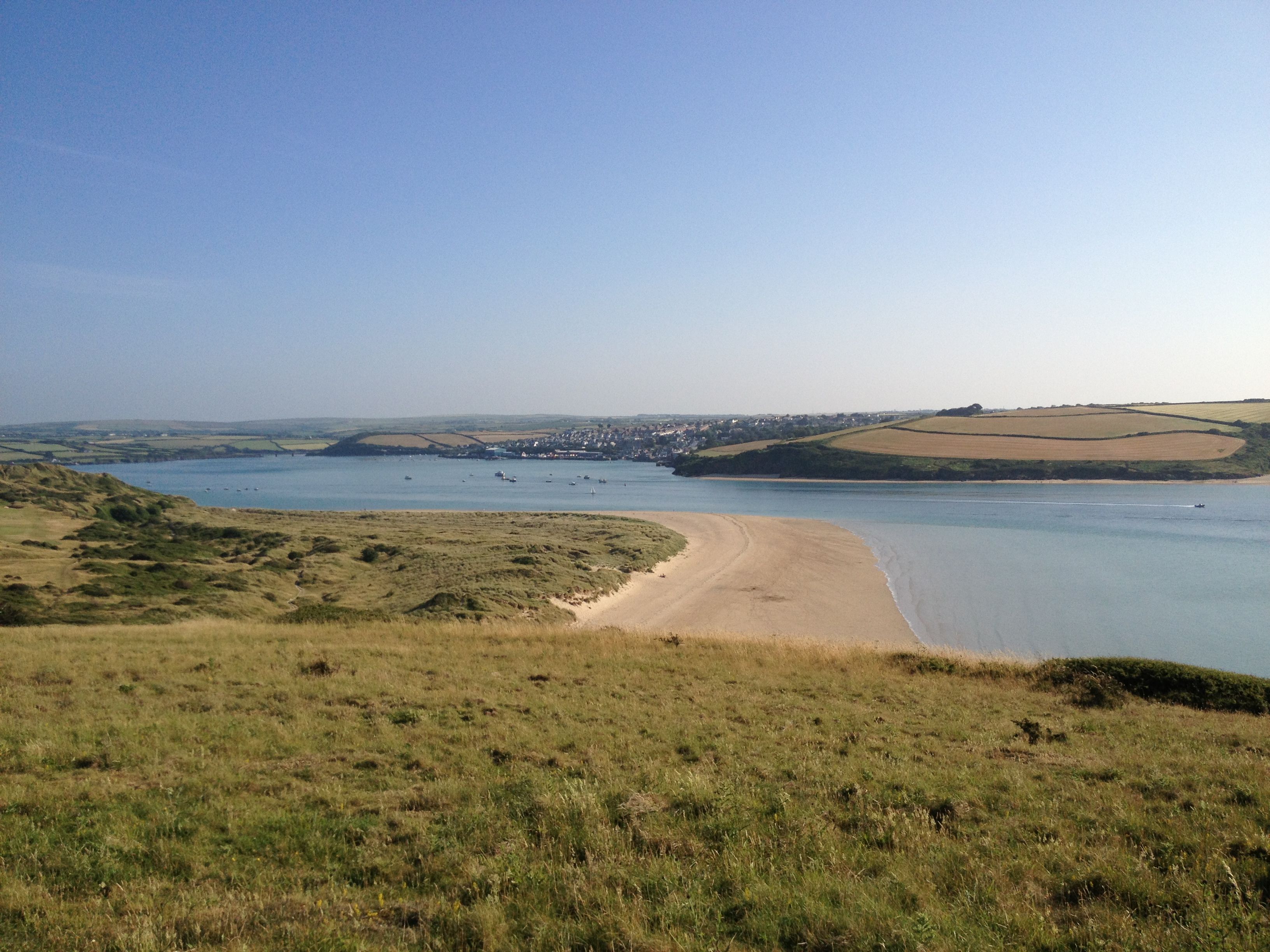 View of a wide river or estuary with grassy hills in the foreground, sandy shoreline, and fields on the opposite bank under a clear blue sky.