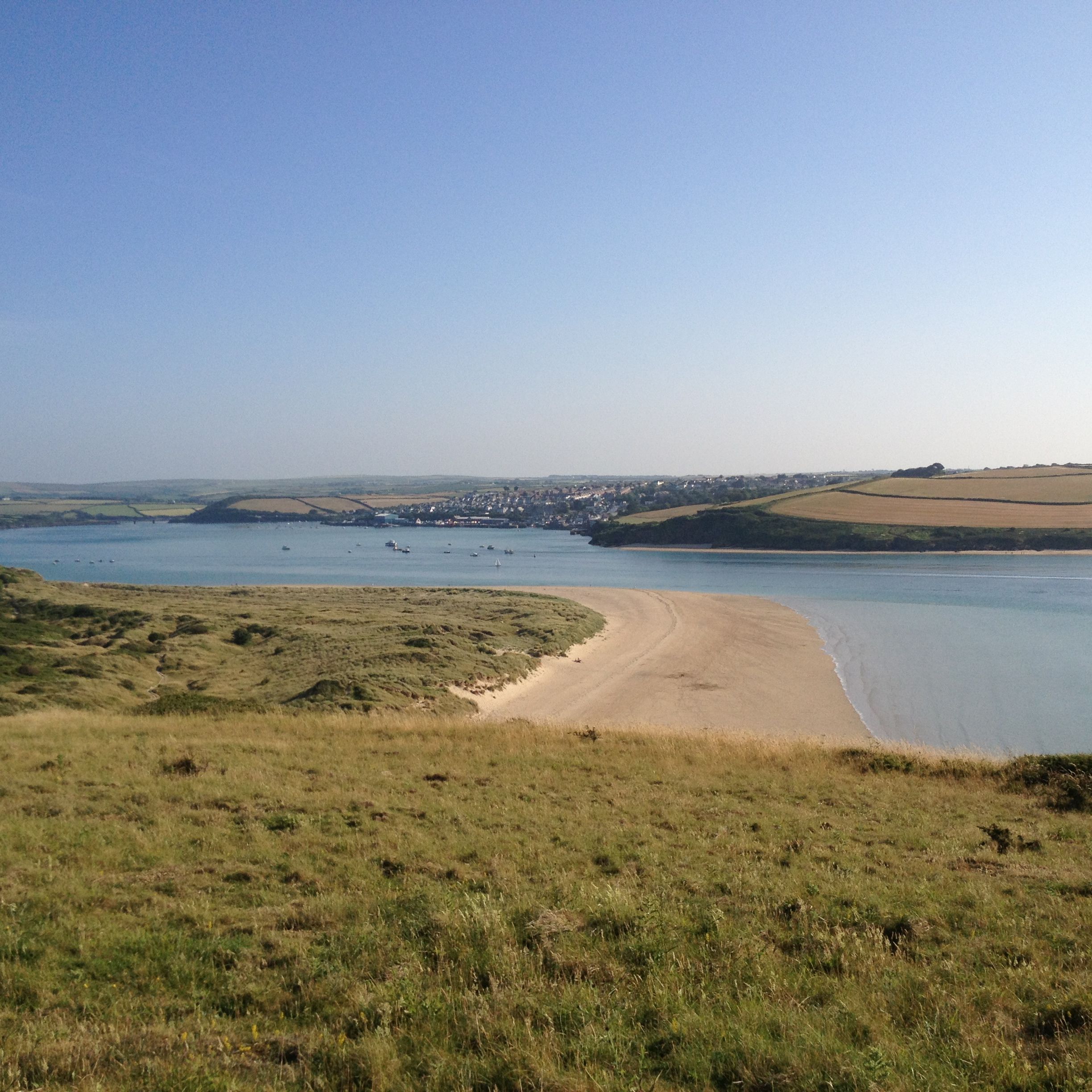 View of a wide river or estuary with grassy hills in the foreground, sandy shoreline, and fields on the opposite bank under a clear blue sky.