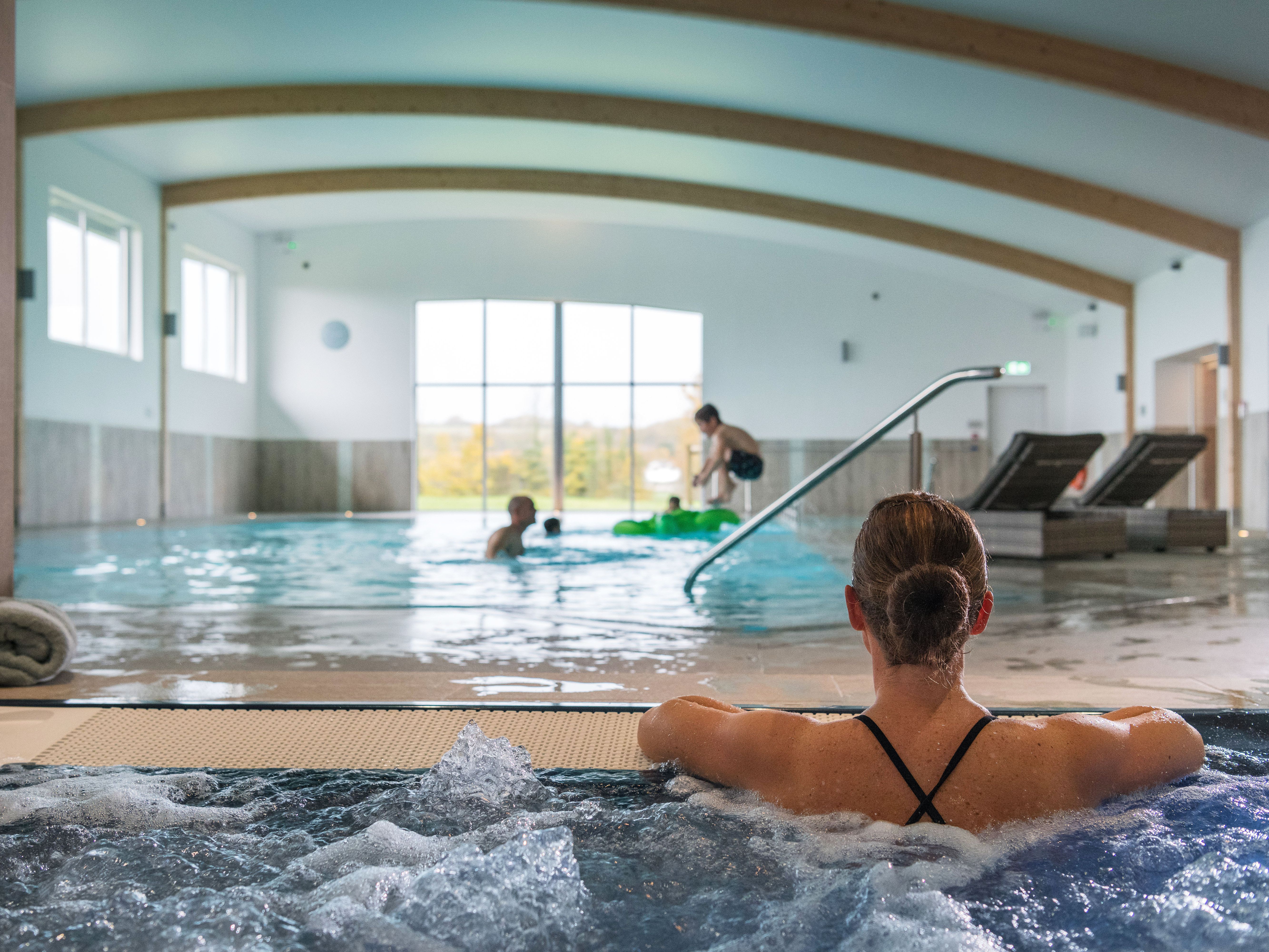 Woman relaxing in a hot tub with people swimming in an indoor pool in the background.