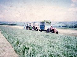 Vintage photo of tractors and farm machinery working in a grassy field with a scenic rural landscape in the background.