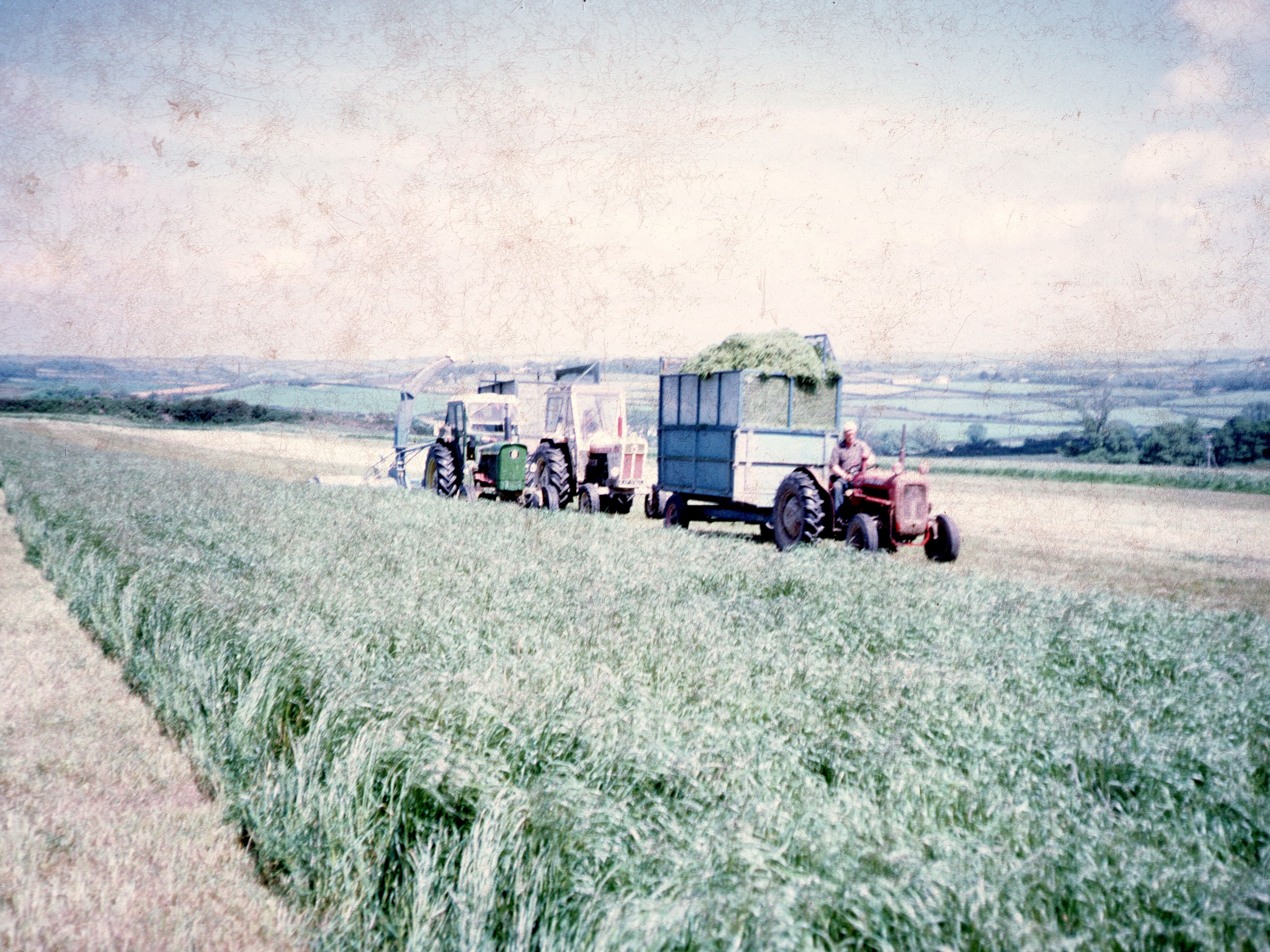 Vintage photo of tractors and farm machinery working in a grassy field with a scenic rural landscape in the background.