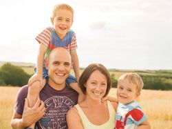 Smiling family of four posing together outdoors in a sunny field.