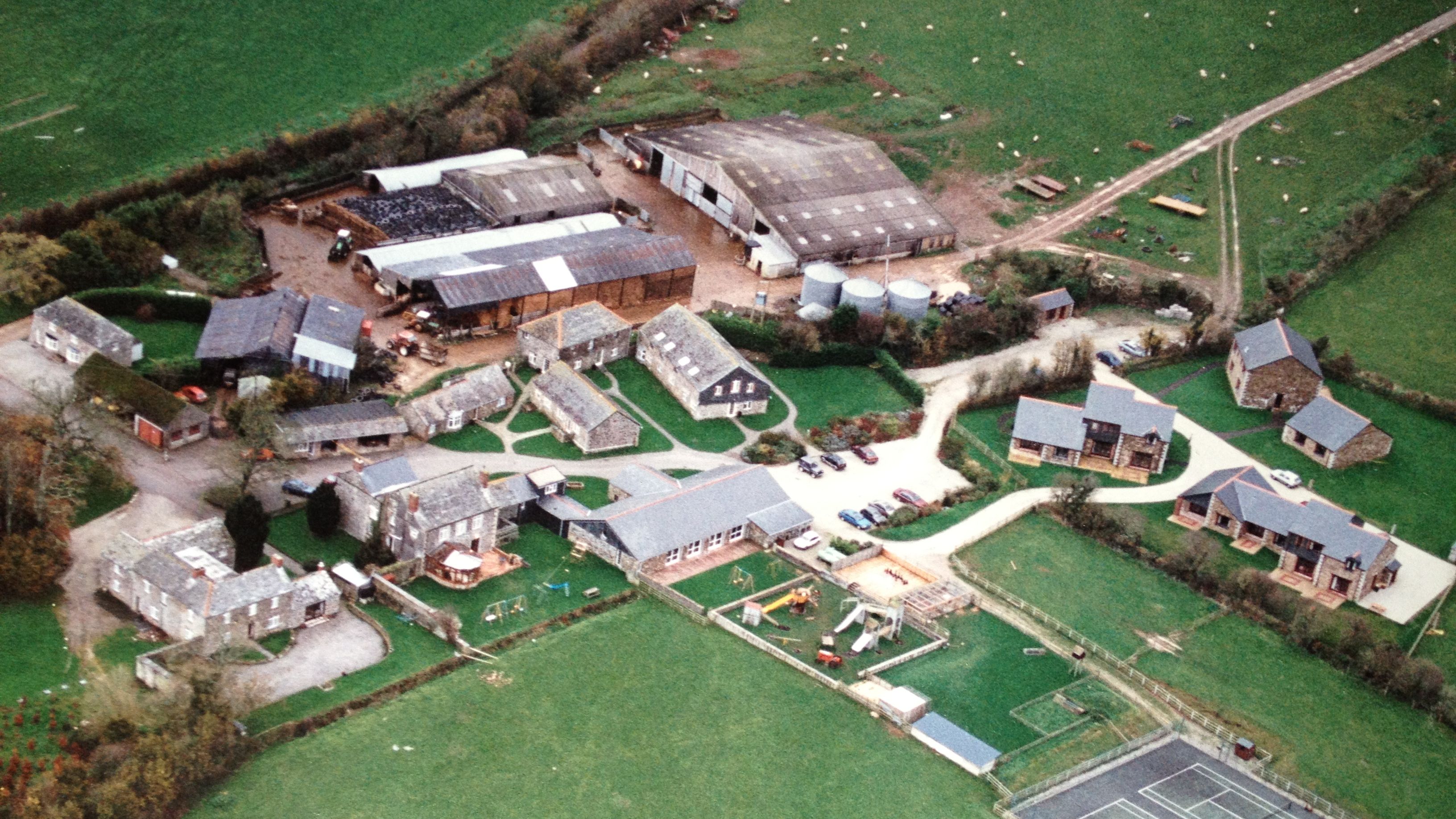 Aerial view of a rural farm complex with several stone buildings, modern barns, agricultural structures, a playground, parking area with cars, tennis courts, and surrounding green fields.
